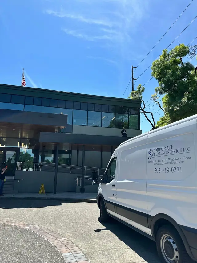 Corporate Cleaning Service van parked outside a glass fronted commercial building, with a crew member cleaning windows from the upper balcony
