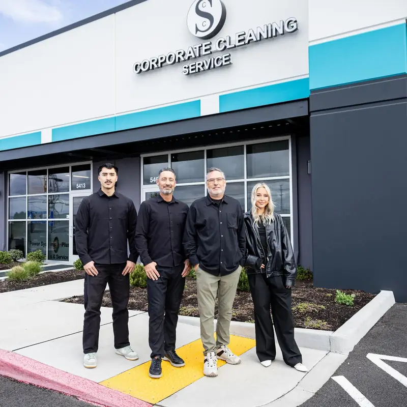 Four members of the Corporate Cleaning Service team standing outside the company's Hillsboro storefront, under its illuminated sign